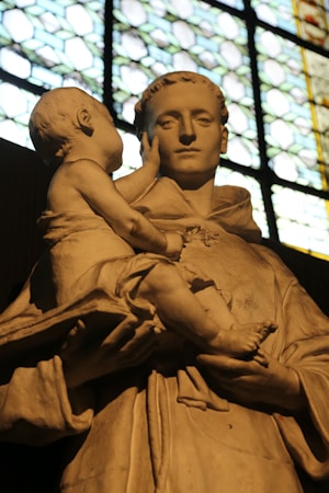 A stone sculpture depicting a serene adult figure holding a child. The adult has a calm expression, while the child, looking up at the adult, gently touches the adult's face. The background includes a stained glass window, allowing light to illuminate the sculpture, highlighting the details and textures.