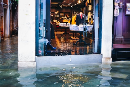 A retail clothing store is partially flooded, with water covering the entrance and forming a reflective surface inside. The store has neatly stacked shirts and brightly colored jackets on display, and mannequins are positioned near the glass front. A person wearing boots is visible inside, standing or seated near the entrance.