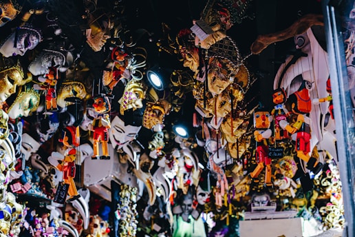 An array of colorful and intricately designed masks and marionette puppets hanging in a shop. The masks feature elaborate decorations and various facial expressions, with some adorned with feathers and glitter. The puppets have a classic design, reminiscent of the famous wooden character Pinocchio, and are suspended on strings.