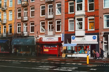 A busy urban street scene featuring a row of brick buildings with multiple windows. Several storefronts are visible, including a coffee shop with a bright red facade and a bicycle shop with blue signage. The street has a designated bike lane, and a couple of people are visible near the entrance of one of the shops.