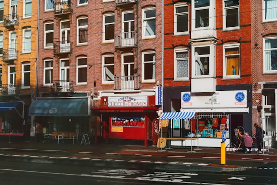 A busy urban street scene featuring a row of brick buildings with multiple windows. Several storefronts are visible, including a coffee shop with a bright red facade and a bicycle shop with blue signage. The street has a designated bike lane, and a couple of people are visible near the entrance of one of the shops.