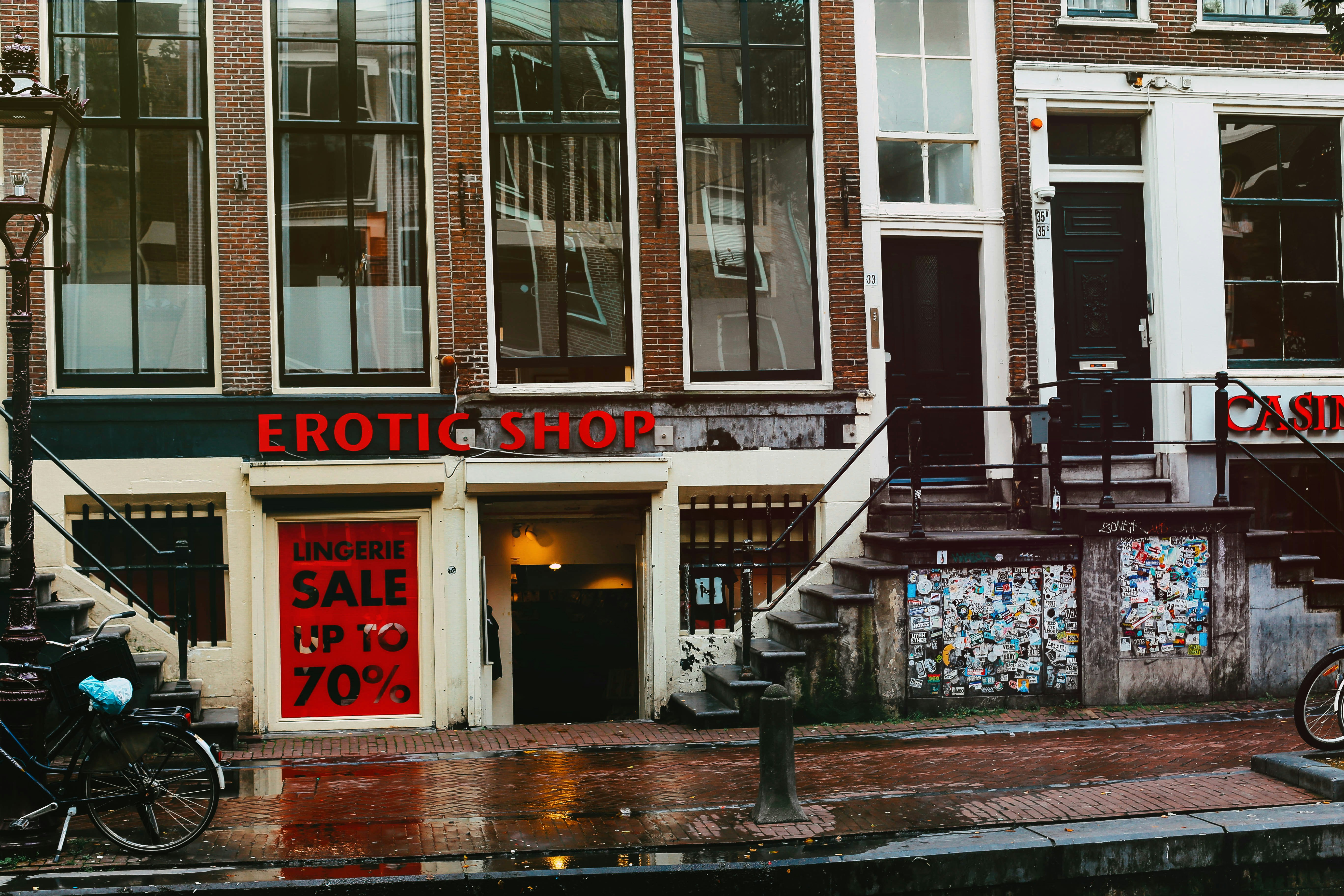 A street view featuring an old brick building with large windows and a shop at the bottom level. The shop has a prominent sign reading 'EROTIC SHOP' in red letters. Below, there is another sign advertising a lingerie sale with discounts up to 70%. Two bikes are parked nearby, and the ground is wet, suggesting recent rain. The building has stairs leading to different entrances and is covered with posters and stickers near the base.
