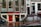 A street view featuring an old brick building with large windows and a shop at the bottom level. The shop has a prominent sign reading 'EROTIC SHOP' in red letters. Below, there is another sign advertising a lingerie sale with discounts up to 70%. Two bikes are parked nearby, and the ground is wet, suggesting recent rain. The building has stairs leading to different entrances and is covered with posters and stickers near the base.