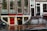 A street view featuring an old brick building with large windows and a shop at the bottom level. The shop has a prominent sign reading 'EROTIC SHOP' in red letters. Below, there is another sign advertising a lingerie sale with discounts up to 70%. Two bikes are parked nearby, and the ground is wet, suggesting recent rain. The building has stairs leading to different entrances and is covered with posters and stickers near the base.