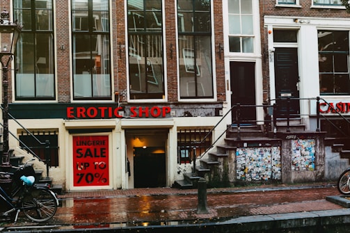 A street view featuring an old brick building with large windows and a shop at the bottom level. The shop has a prominent sign reading 'EROTIC SHOP' in red letters. Below, there is another sign advertising a lingerie sale with discounts up to 70%. Two bikes are parked nearby, and the ground is wet, suggesting recent rain. The building has stairs leading to different entrances and is covered with posters and stickers near the base.