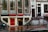 A street view featuring an old brick building with large windows and a shop at the bottom level. The shop has a prominent sign reading 'EROTIC SHOP' in red letters. Below, there is another sign advertising a lingerie sale with discounts up to 70%. Two bikes are parked nearby, and the ground is wet, suggesting recent rain. The building has stairs leading to different entrances and is covered with posters and stickers near the base.