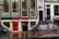 A street view featuring an old brick building with large windows and a shop at the bottom level. The shop has a prominent sign reading 'EROTIC SHOP' in red letters. Below, there is another sign advertising a lingerie sale with discounts up to 70%. Two bikes are parked nearby, and the ground is wet, suggesting recent rain. The building has stairs leading to different entrances and is covered with posters and stickers near the base.
