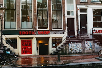 A street view featuring an old brick building with large windows and a shop at the bottom level. The shop has a prominent sign reading 'EROTIC SHOP' in red letters. Below, there is another sign advertising a lingerie sale with discounts up to 70%. Two bikes are parked nearby, and the ground is wet, suggesting recent rain. The building has stairs leading to different entrances and is covered with posters and stickers near the base.