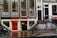 A street view featuring an old brick building with large windows and a shop at the bottom level. The shop has a prominent sign reading 'EROTIC SHOP' in red letters. Below, there is another sign advertising a lingerie sale with discounts up to 70%. Two bikes are parked nearby, and the ground is wet, suggesting recent rain. The building has stairs leading to different entrances and is covered with posters and stickers near the base.