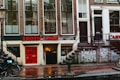 A street view featuring an old brick building with large windows and a shop at the bottom level. The shop has a prominent sign reading 'EROTIC SHOP' in red letters. Below, there is another sign advertising a lingerie sale with discounts up to 70%. Two bikes are parked nearby, and the ground is wet, suggesting recent rain. The building has stairs leading to different entrances and is covered with posters and stickers near the base.