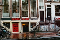 A street view featuring an old brick building with large windows and a shop at the bottom level. The shop has a prominent sign reading 'EROTIC SHOP' in red letters. Below, there is another sign advertising a lingerie sale with discounts up to 70%. Two bikes are parked nearby, and the ground is wet, suggesting recent rain. The building has stairs leading to different entrances and is covered with posters and stickers near the base.