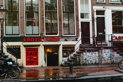 A street view featuring an old brick building with large windows and a shop at the bottom level. The shop has a prominent sign reading 'EROTIC SHOP' in red letters. Below, there is another sign advertising a lingerie sale with discounts up to 70%. Two bikes are parked nearby, and the ground is wet, suggesting recent rain. The building has stairs leading to different entrances and is covered with posters and stickers near the base.