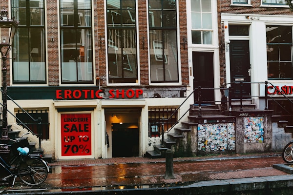 A street view featuring an old brick building with large windows and a shop at the bottom level. The shop has a prominent sign reading 'EROTIC SHOP' in red letters. Below, there is another sign advertising a lingerie sale with discounts up to 70%. Two bikes are parked nearby, and the ground is wet, suggesting recent rain. The building has stairs leading to different entrances and is covered with posters and stickers near the base.