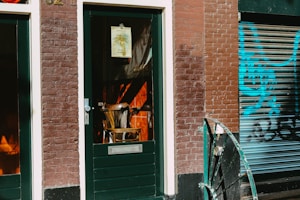 A rustic door with a glass panel is adjacent to a brick wall. Through the glass, a wooden chair is visible inside. The exterior features brown and white bricks, with a poster fixed to the door. Beside the door, a shutter shows graffiti in vibrant blue and black, partially visible. The dark green frame and railings complement the brick façade.