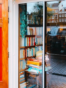 A cozy bookshop view through a window shows shelves filled with books of various sizes. A small wooden table holds stacked board games, including a visible box of Scrabble. Reflections of the street outside, including cars and buildings, can be seen on the glass. A decorative plant hangs, adding a touch of greenery.