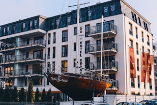 A modern multi-story hotel building with numerous balconies, set against a clear sky. In front of the building is a large boat on display, with the name 'Hotel Gdańsk' visible on the hull. The structure and design suggest a contemporary and clean architectural style.