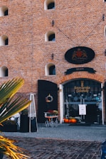 A brick building with arched windows features a prominent entrance labeled 'Brovarnia Gdańsk.' The entrance has a black oval sign with a crest, and there is a neatly arranged outdoor decor including a heater, plants, a decorative cart, and several pumpkins. The building is adorned with a sign indicating the year 1690, suggesting historical significance.