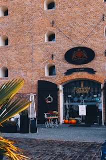 A brick building with arched windows features a prominent entrance labeled 'Brovarnia Gdańsk.' The entrance has a black oval sign with a crest, and there is a neatly arranged outdoor decor including a heater, plants, a decorative cart, and several pumpkins. The building is adorned with a sign indicating the year 1690, suggesting historical significance.