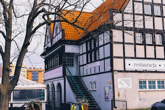 A traditional European-style building with a distinctive orange-tiled roof and wooden beam accents is nestled behind a bare tree. A staircase leads up the side of the building, and several signs are visible, indicating it is a restaurant and hotel. Surrounding the area are other modern buildings, adding contrast to the scene.