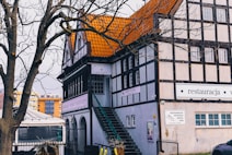 A traditional European-style building with a distinctive orange-tiled roof and wooden beam accents is nestled behind a bare tree. A staircase leads up the side of the building, and several signs are visible, indicating it is a restaurant and hotel. Surrounding the area are other modern buildings, adding contrast to the scene.