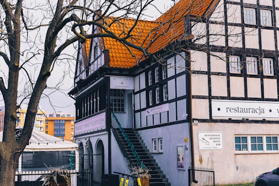 A traditional European-style building with a distinctive orange-tiled roof and wooden beam accents is nestled behind a bare tree. A staircase leads up the side of the building, and several signs are visible, indicating it is a restaurant and hotel. Surrounding the area are other modern buildings, adding contrast to the scene.