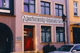 A street view of a building with a sign reading 'Apartamenty Gdański Gród'. The facade is painted pink with multiple rectangular windows. There is a red door at the entrance with decorative glasswork. A lamp post is visible on the right side, and part of a car is parked nearby. The neighboring buildings are also visible, painted in different colors.