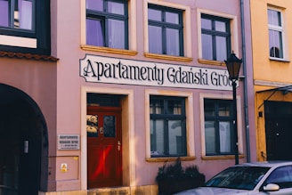 A street view of a building with a sign reading 'Apartamenty Gdański Gród'. The facade is painted pink with multiple rectangular windows. There is a red door at the entrance with decorative glasswork. A lamp post is visible on the right side, and part of a car is parked nearby. The neighboring buildings are also visible, painted in different colors.