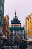 A narrow street lined with colorful buildings leads to a historic architectural structure with ornate details and a tower. People are walking along the street, creating a lively atmosphere. The architecture features classic design elements and statues, indicating a rich cultural heritage.