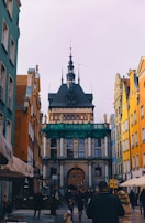 A narrow street lined with colorful buildings leads to a historic architectural structure with ornate details and a tower. People are walking along the street, creating a lively atmosphere. The architecture features classic design elements and statues, indicating a rich cultural heritage.