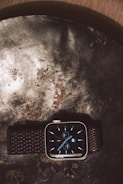 A smartwatch with a dark woven strap displayed on a textured metallic surface, featuring a digital analog-style clock face with blue hour markers and subdials.