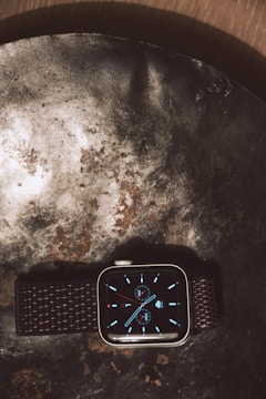A smartwatch with a dark woven strap displayed on a textured metallic surface, featuring a digital analog-style clock face with blue hour markers and subdials.