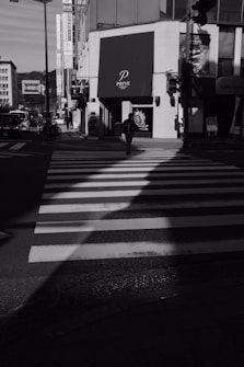 A black and white photograph of an urban intersection featuring a zebra crossing leading toward a modern building with a prominent sign that reads 'PRIVE'. A lone figure walks across the crosswalk surrounded by storefronts, streetlights, and a minimal amount of vehicular traffic. Harsh shadows and bright highlights create a contrast across the scene.