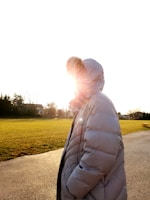 A person wearing a large puffer jacket with a fur-trimmed hood, standing in an open park space during daylight. The sun is shining brightly, partially obscuring the view with lens flare and creating a warm glow around the figure. The background features green grass and distant tree-lined residential homes.