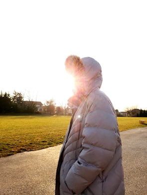 A person wearing a large puffer jacket with a fur-trimmed hood, standing in an open park space during daylight. The sun is shining brightly, partially obscuring the view with lens flare and creating a warm glow around the figure. The background features green grass and distant tree-lined residential homes.