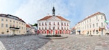 A historic town square features an ornate building with a red roof and white façade adorned with flower boxes, surrounded by other classical-style buildings. The square is paved with cobblestones and includes a fountain at its center.