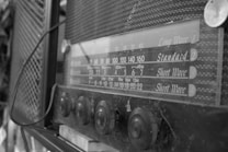 A vintage radio with a front panel displaying frequency ranges, accompanied by various control knobs. The radio has a mesh pattern on the casing and appears to be from an earlier era, indicated by its design and labeling.