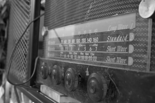 A vintage radio with a front panel displaying frequency ranges, accompanied by various control knobs. The radio has a mesh pattern on the casing and appears to be from an earlier era, indicated by its design and labeling.