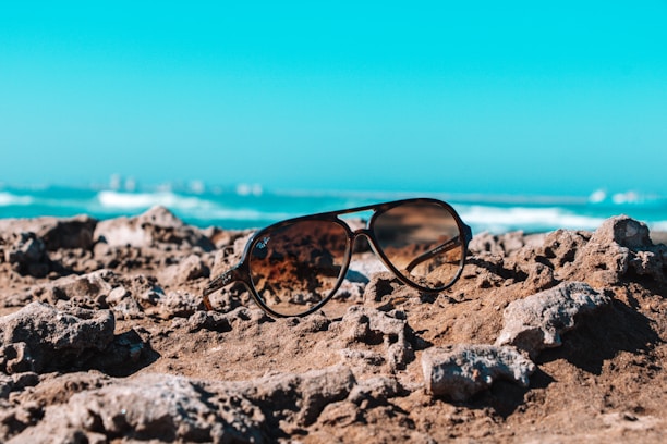 A pair of sleek sunglasses placed on a sunny beach towel with the ocean blurred in the background.