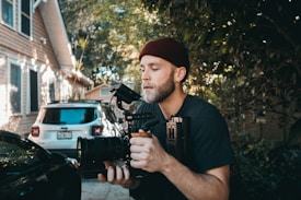 A person wearing a beanie is holding and operating a professional video camera outdoors. They are standing beside a house and a parked car in a driveway, surrounded by greenery. The lighting suggests a sunny day with dappled sunlight filtering through the trees.