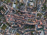 An aerial view of a city layout with densely arranged buildings, roads, and intersections. The image captures a mix of residential and commercial structures with red-tiled roofs and modern buildings, surrounded by green spaces and tree-lined streets.