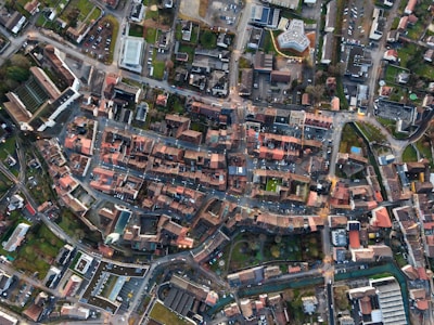 An aerial view of a city layout with densely arranged buildings, roads, and intersections. The image captures a mix of residential and commercial structures with red-tiled roofs and modern buildings, surrounded by green spaces and tree-lined streets.