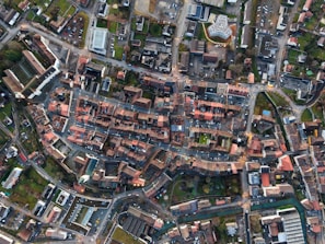 An aerial view of a city layout with densely arranged buildings, roads, and intersections. The image captures a mix of residential and commercial structures with red-tiled roofs and modern buildings, surrounded by green spaces and tree-lined streets.