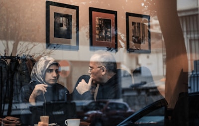 Two people are sitting at a table in a cozy café, engaged in a thoughtful conversation. The scene is viewed through a reflective window, which shows street reflections alongside the interior of the café. Three framed pictures are visible on the café wall behind them.