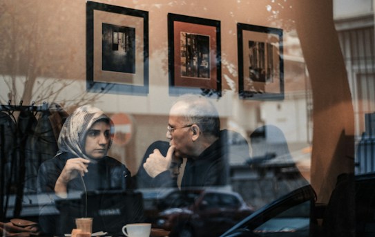 Two people are sitting at a table in a cozy caf&eacute;, engaged in a thoughtful conversation. The scene is viewed through a reflective window, which shows street reflections alongside the interior of the caf&eacute;. Three framed pictures are visible on the caf&eacute; wall behind them.