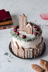 A decorated cake featuring gingerbread cookies shaped like houses, topped with sprigs of greenery and vibrant red cherries. The cake is on a metal plate, surrounded by a minimalist, light-toned setting. In the background, there are stacked books with red and brown covers and a glass with a pink liquid.
