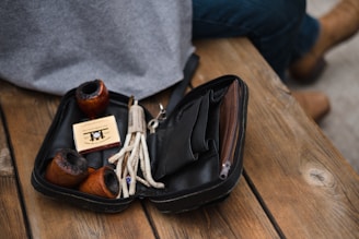 A collection of smoking pipes rests inside a black leather pouch. The pouch contains three wooden pipes, a small box, and pipe cleaners. These items are arranged on a wooden surface, possibly a bench, with a person wearing jeans and brown shoes nearby.