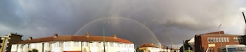 A double rainbow arches over a residential area, where the houses have a uniform style with red-tiled roofs and white walls. Dark clouds loom in the sky, contrasting with a small patch of blue, while the rainbows add vibrant colors to the scene. An older-style bus is partially visible on the right side of the image.