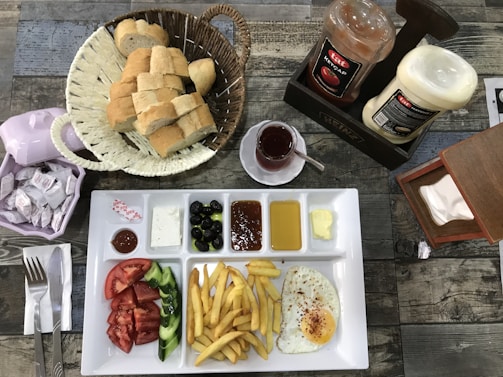 A breakfast spread with a white sectioned plate containing fries, a sunny-side-up egg, sliced tomatoes, cucumbers, black olives, jam, butter, white cheese, and various sauces. Accompanying the meal are a basket of sliced bread, packets of cream, a small cup of tea, and bottles of ketchup and mayonnaise. The table is set with a knife and fork on a folded napkin.