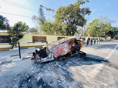 A burnt-out, overturned car lies on a road, surrounded by charred debris. The setting is a tree-lined street with a fence and a yellow wall adorned with murals in the background. Nearby, a few people are observing the scene.