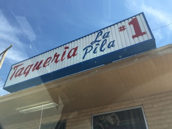 A storefront with a large sign on the roof reading 'Taqueria La Pila #1' in red and blue letters. The building has a brick facade and there is a clear blue sky in the background.